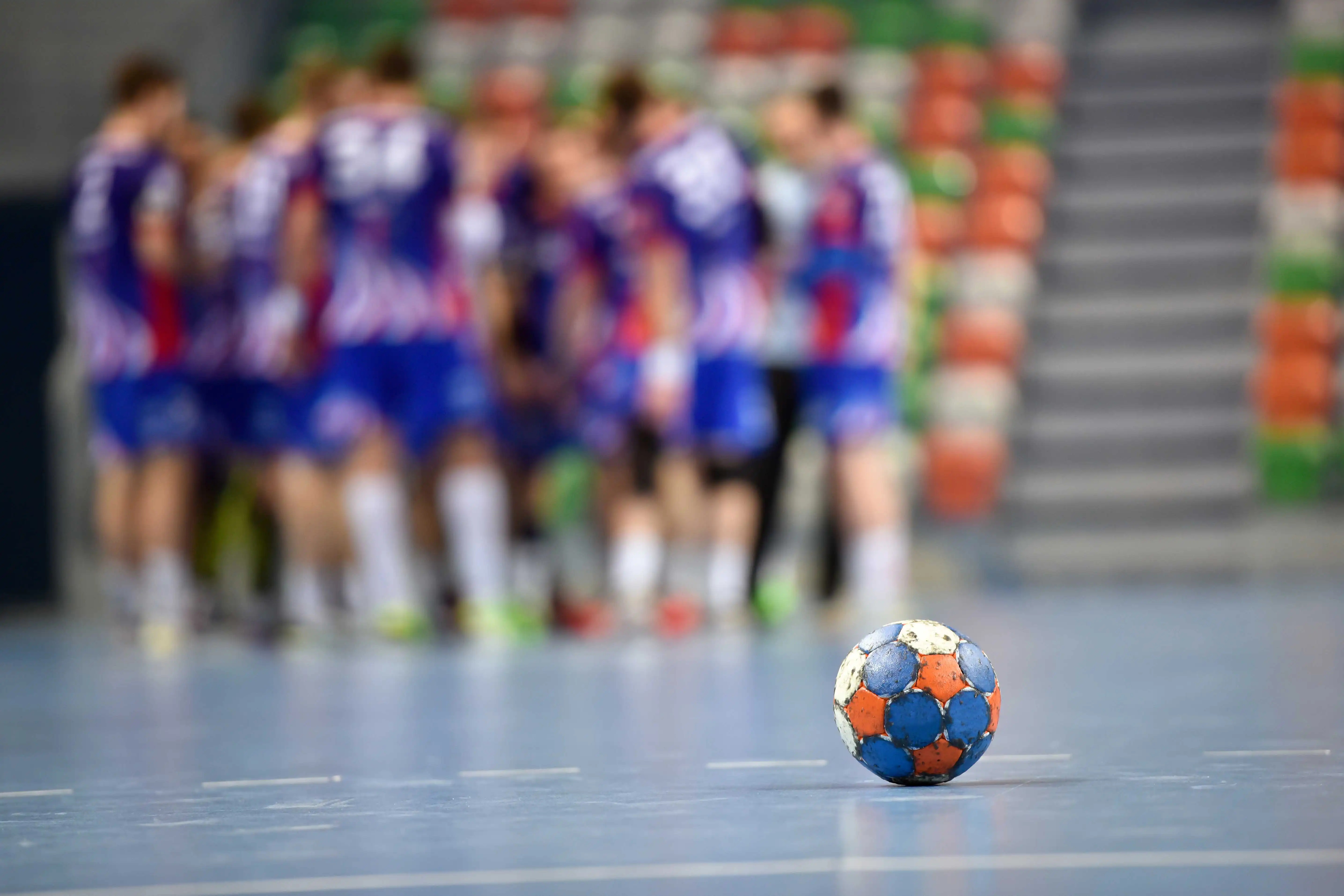 En närbild av en handboll som ligger ensam på ett golv i en idrottshall. I bakgrunden syns en grupp spelare i blå, röda och vita matchdräkter som står samlade i en ring.